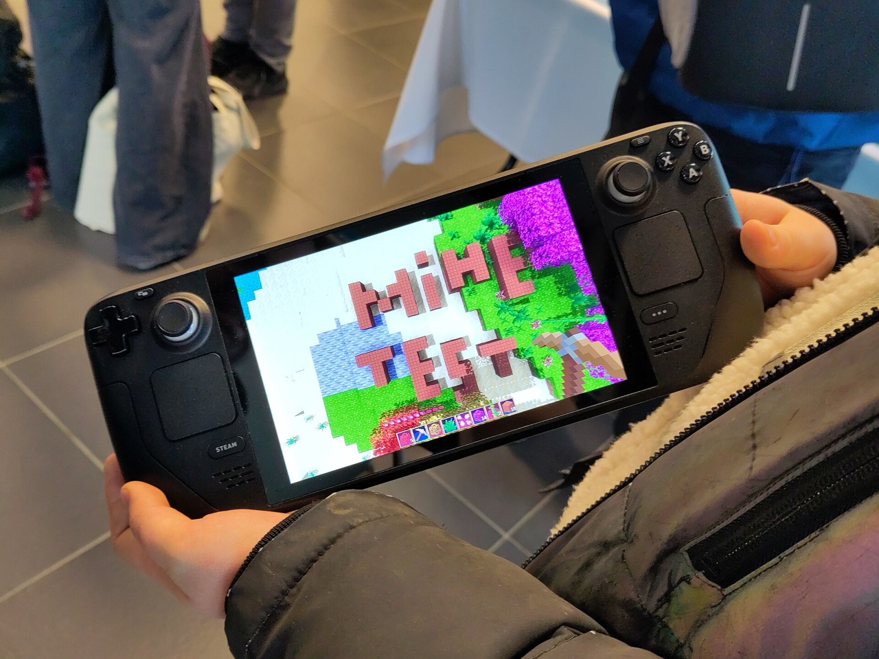 A Steam Deck held in both hands and being played, thumbs on the sticks, at a crowded conference